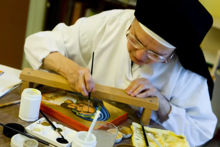 Religieuse âgée concentrée peignant une icône religieuse sur bois avec pinceau et palette de couleurs.