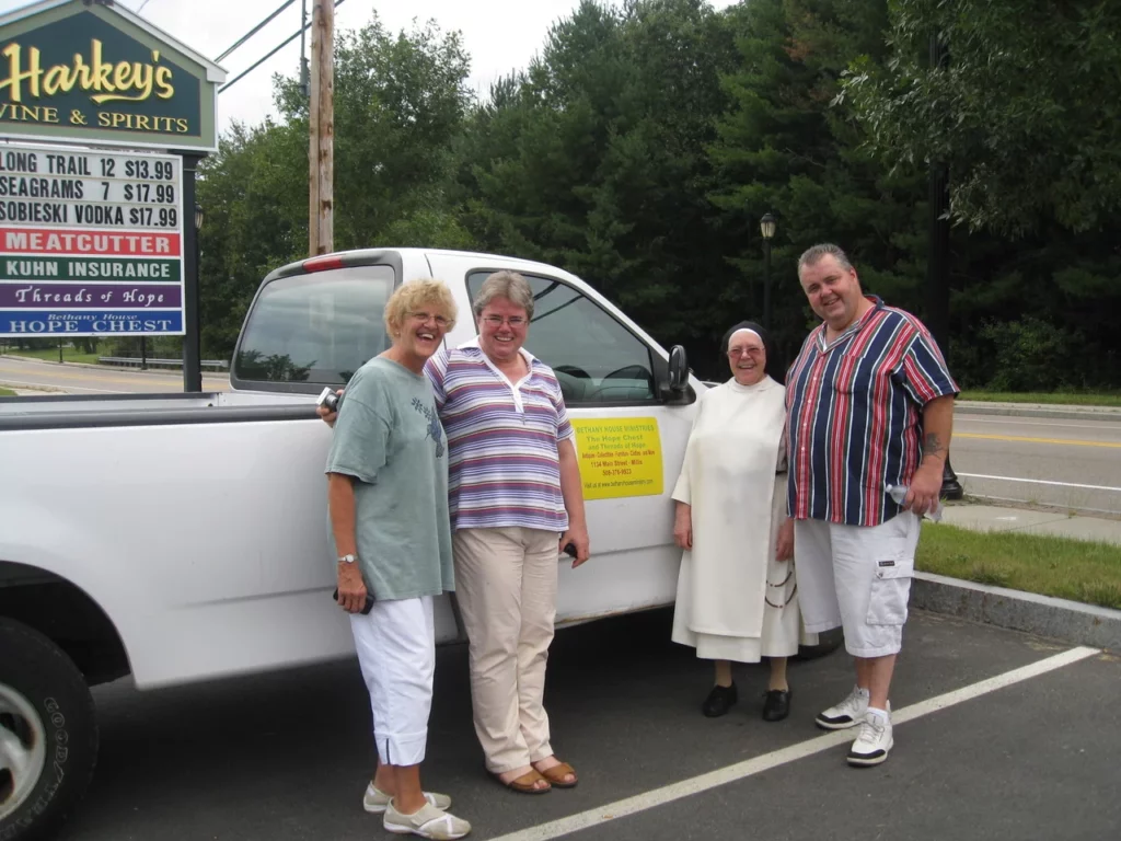 Quatre personnes, dont une nonne, posant devant une camionnette blanche et un panneau publicitaire pour Harkey's Wine & Spirits et Bethany House Ministries.
