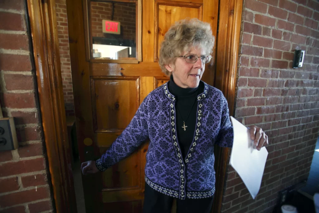 Femme âgée en pull violet à motif, lunettes et pendentif croix, tenant un document près d'une porte en bois avec un mur de briques et un panneau "EXIT".