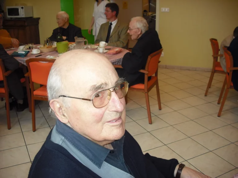 Vieil homme à lunettes en gros plan, regardant vers la droite, lors d'un moment de convivialité avec d'autres personnes âgées dans une résidence.