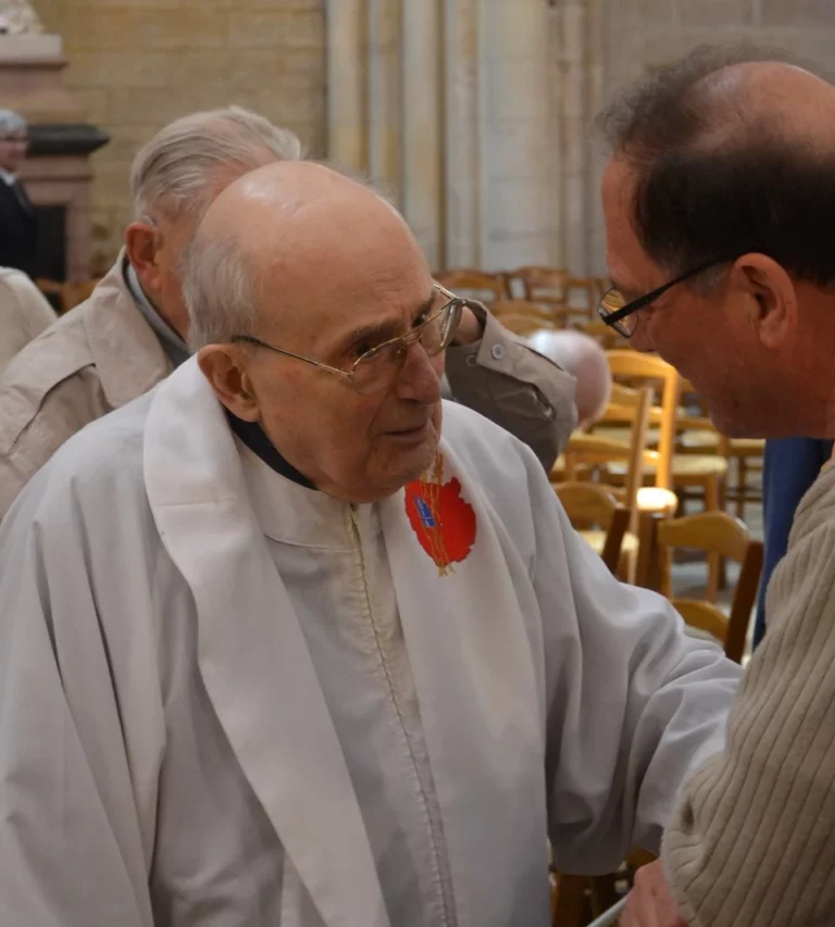 Deux hommes âgés, dont l'un en aube blanche avec un emblème religieux rouge, discutent attentivement dans une église.