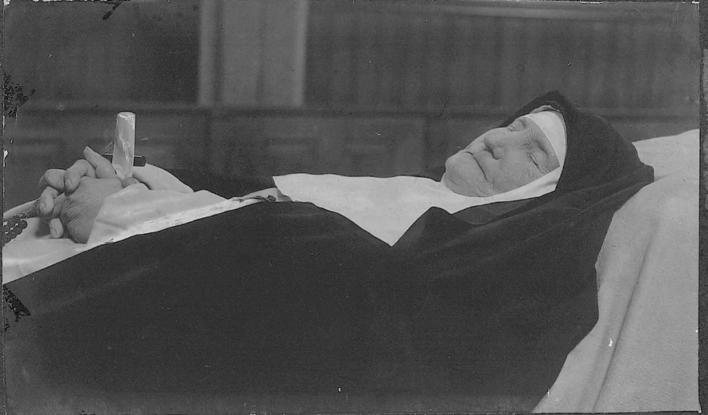 Photo en noir et blanc d'une religieuse âgée décédée, vêtue de son habit, les mains jointes sur la poitrine tenant un crucifix et un chapelet.
