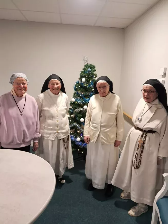 Quatre femmes âgées, dont trois religieuses en habit blanc et voile noir, souriantes devant un sapin de Noël illuminé.